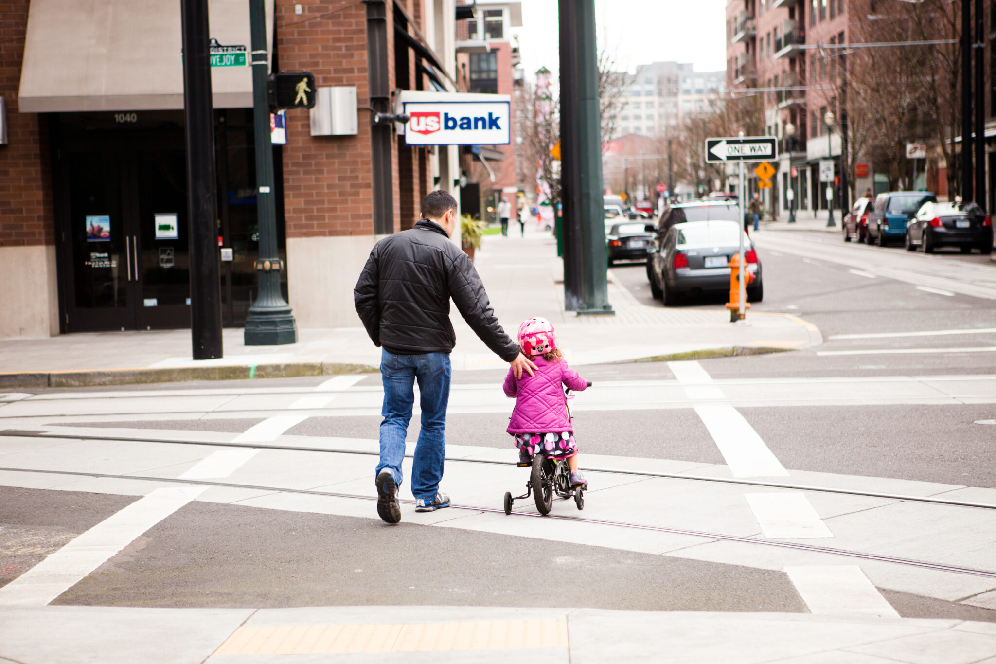 "Portland, Oregon, USA - February 12, 2012: A man helps his daughter across the street as she rides her bicycle with training wheels in the Pearl district of Portland."