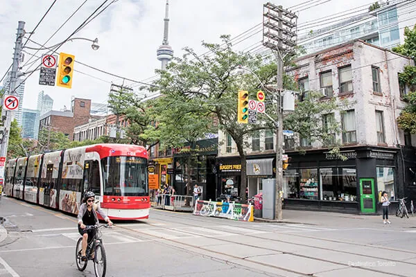 Toronto street scene on King Street West with streetcar, a woman bicycling and storefronts with people walking by. Credit Destination Toronto